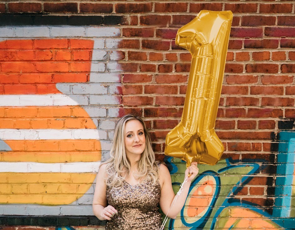 blonde female holding number 1 balloon in front of graffiti wall to celebrate anniversary