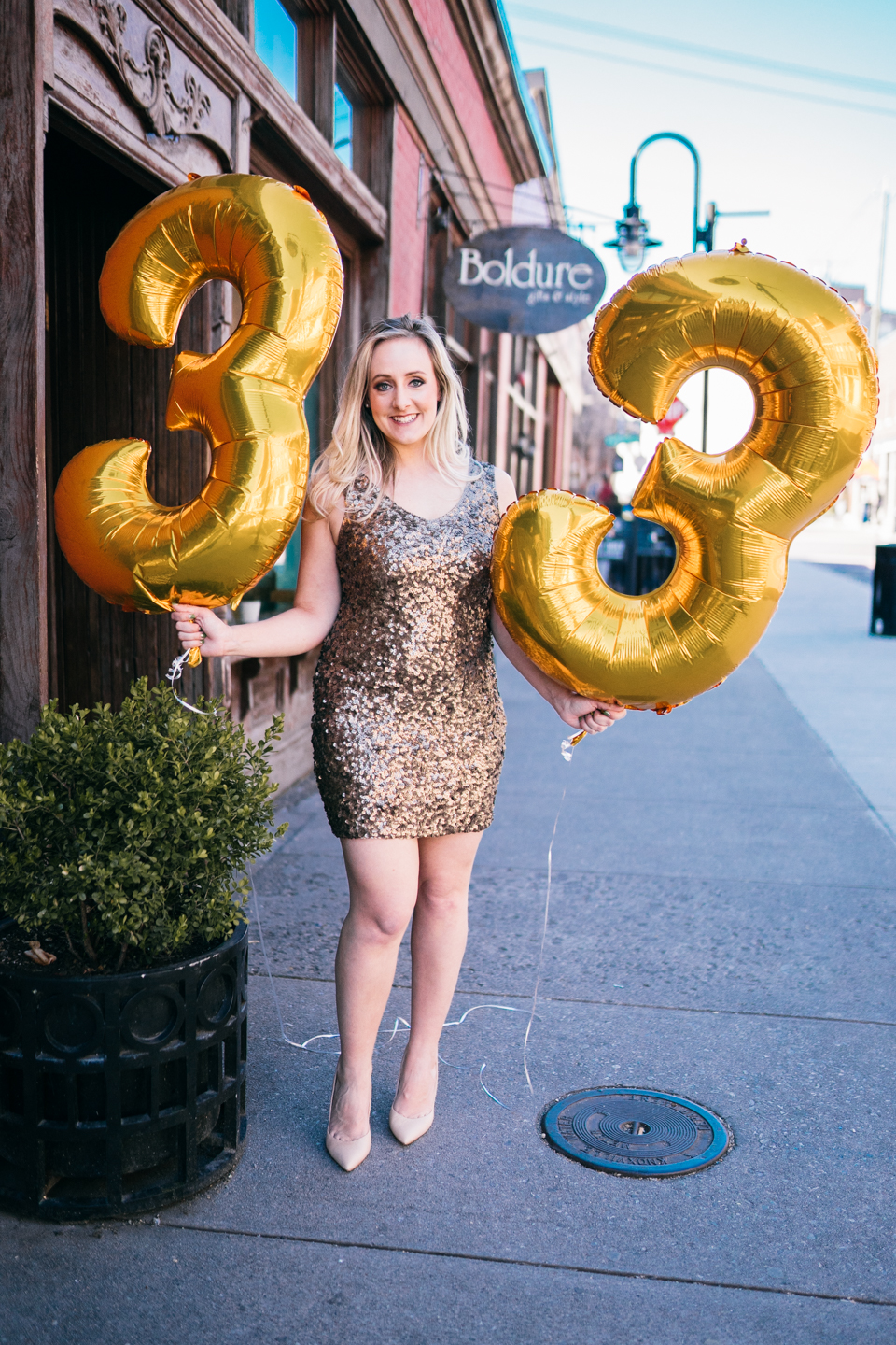 woman in gold sequin party dress holding two large foil balloons that display 33